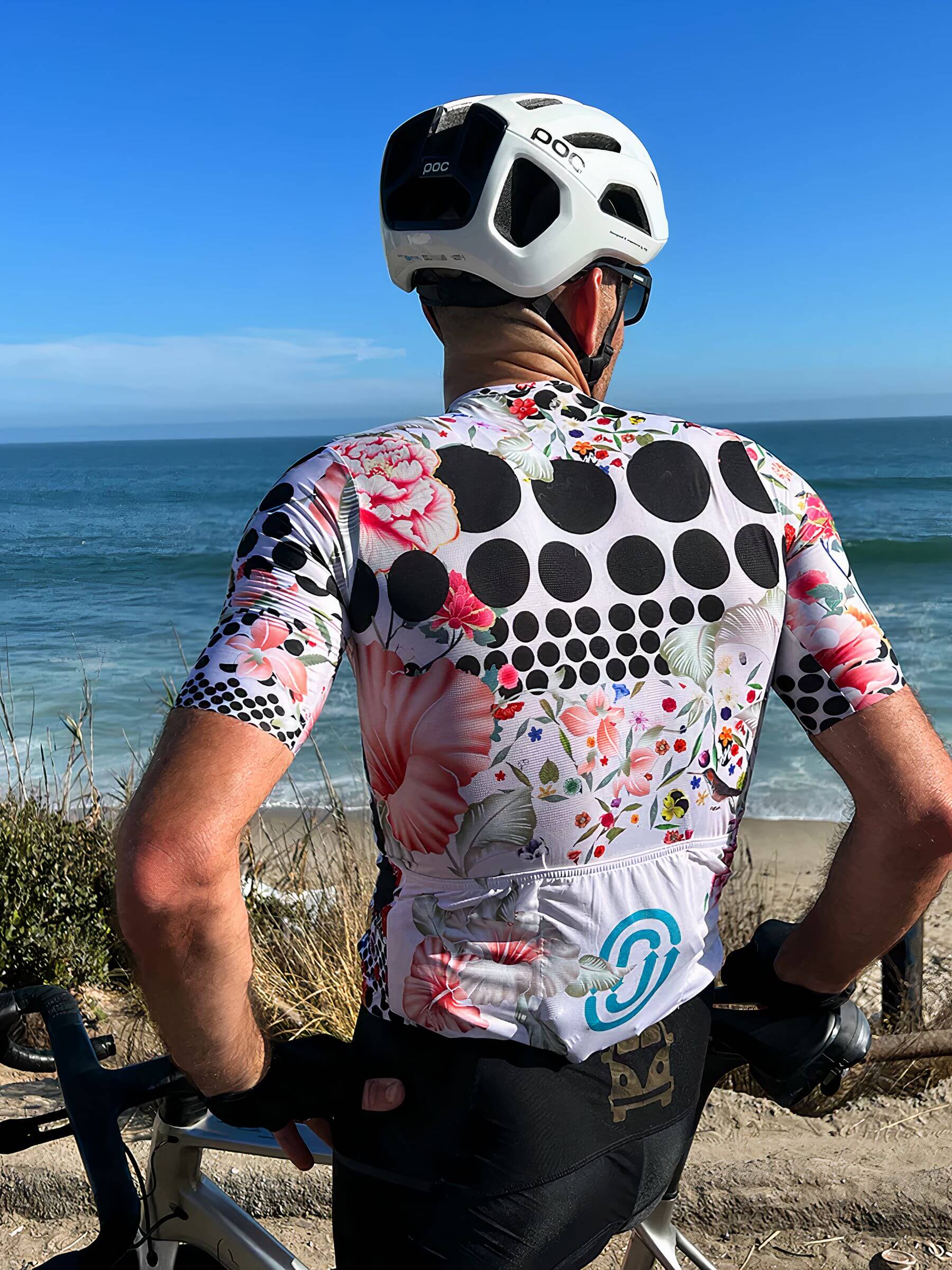 Man wearing Ostroy Omloop White Jersey - Men's with floral and polka dot design standing by the ocean with bike handlebars visible