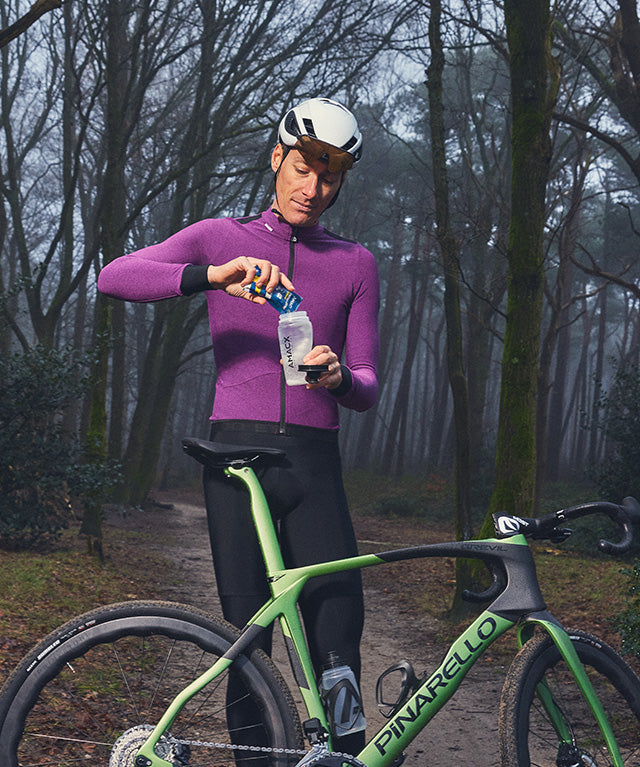Man in cycling gear pouring Amacx Energy Drink On The Go into a bottle beside a green Pinarello bike on forest trail