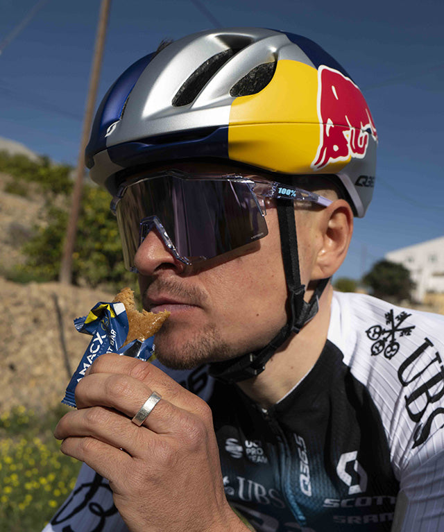 Cyclist wearing helmet and sunglasses eating an Amacx Energy Oat Bar Lemon outdoors on a sunny day