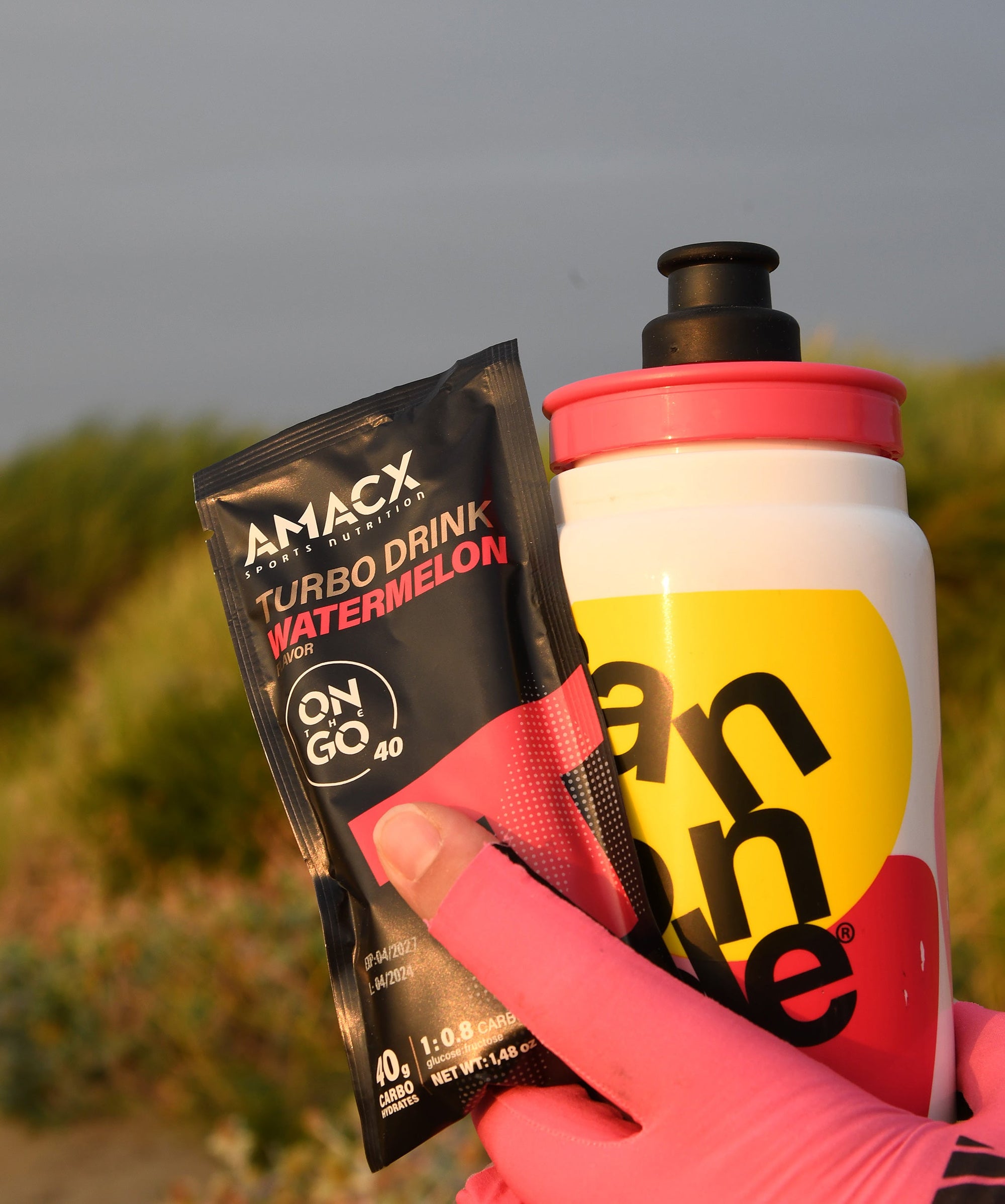 Hand holding Amacx Turbo Drink On The Go Watermelon packet and colorful sports water bottle outdoors.