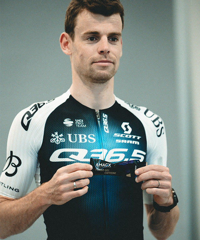 Man in cycling gear holding Amacx Turbo Gel Cassis Caffeine energy gel in hands against neutral background