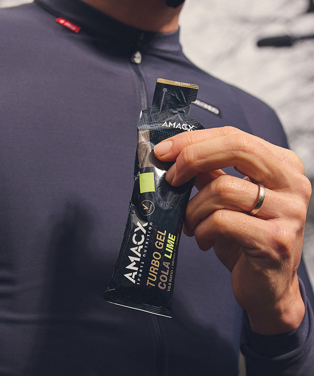 Hand holding a black Amacx Turbo Gel Cola Lime energy gel packet against a person wearing a dark long-sleeve shirt
