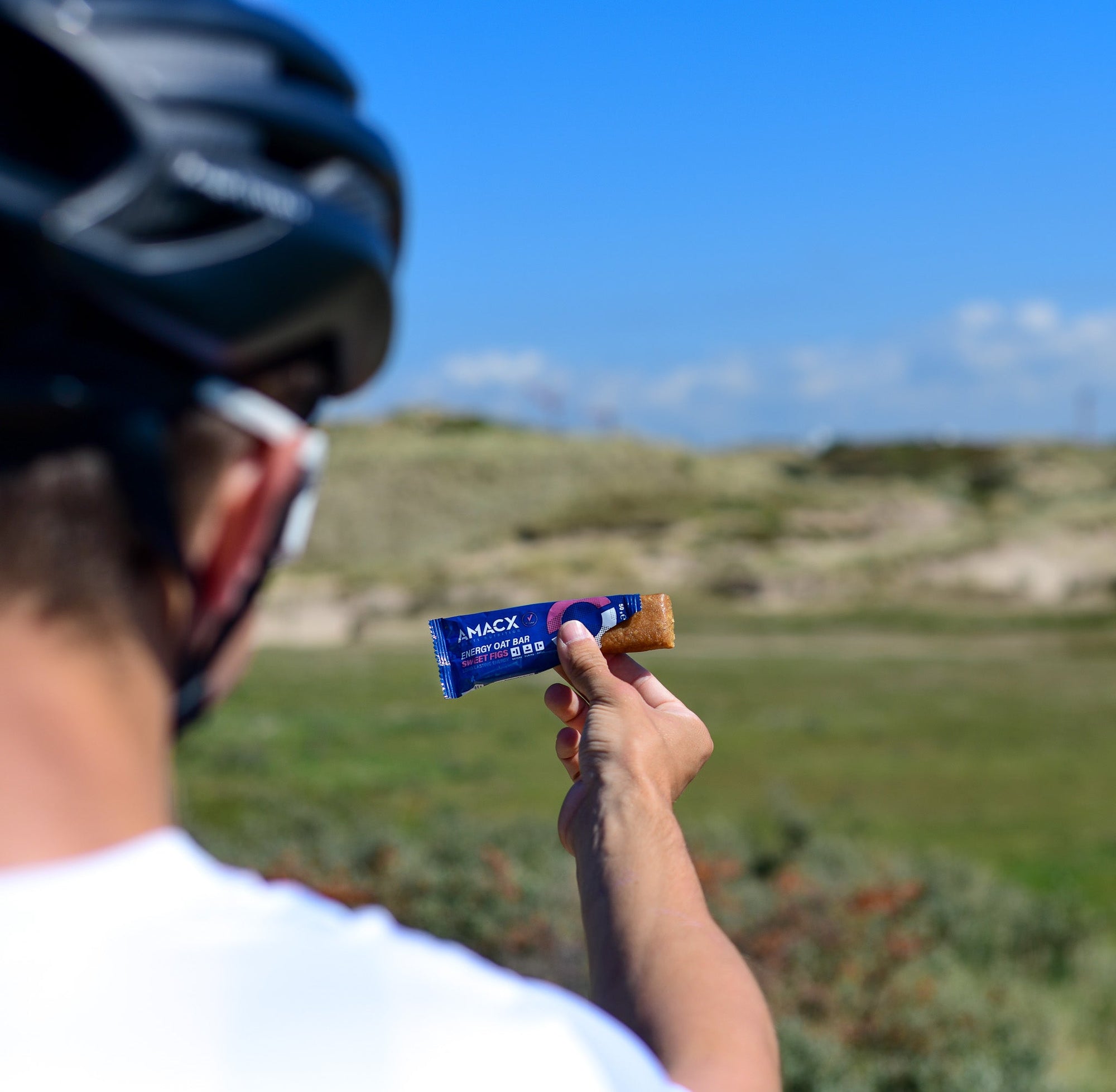 Person wearing a helmet holding an Amacx Energy Oat Bar Sweet Figs outdoors against a clear blue sky.