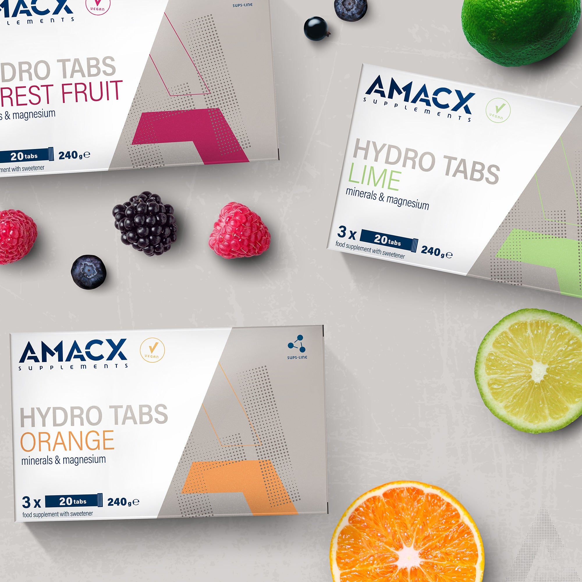 Boxes of Amacx Hydro Tabs Forest Fruit along with lime and orange flavors, fresh berries, lime, and orange slices on gray surface