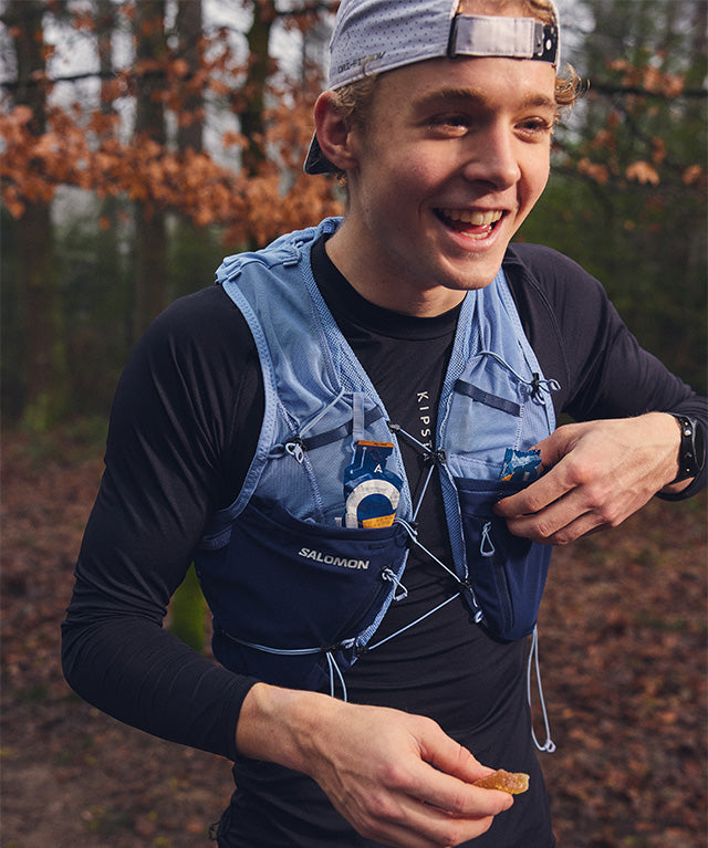 Man wearing a blue running vest holding Amacx Drink Gel Raspberry during an outdoor forest run