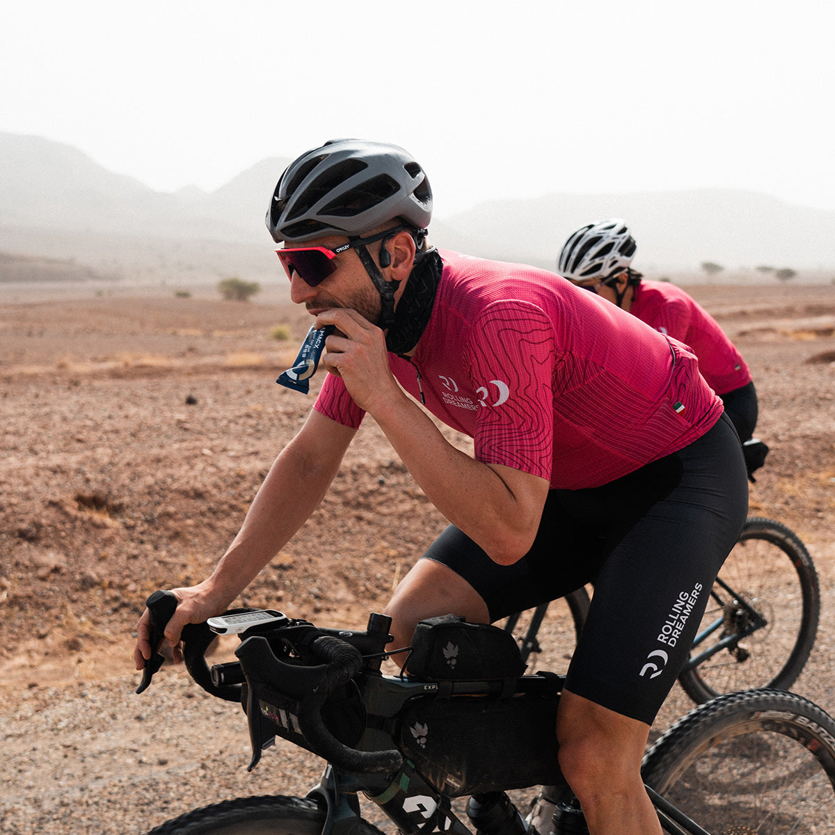 Cyclist wearing a helmet and sunglasses eating an Amacx Energy Oat Bar Coconut while riding a bike outdoors.