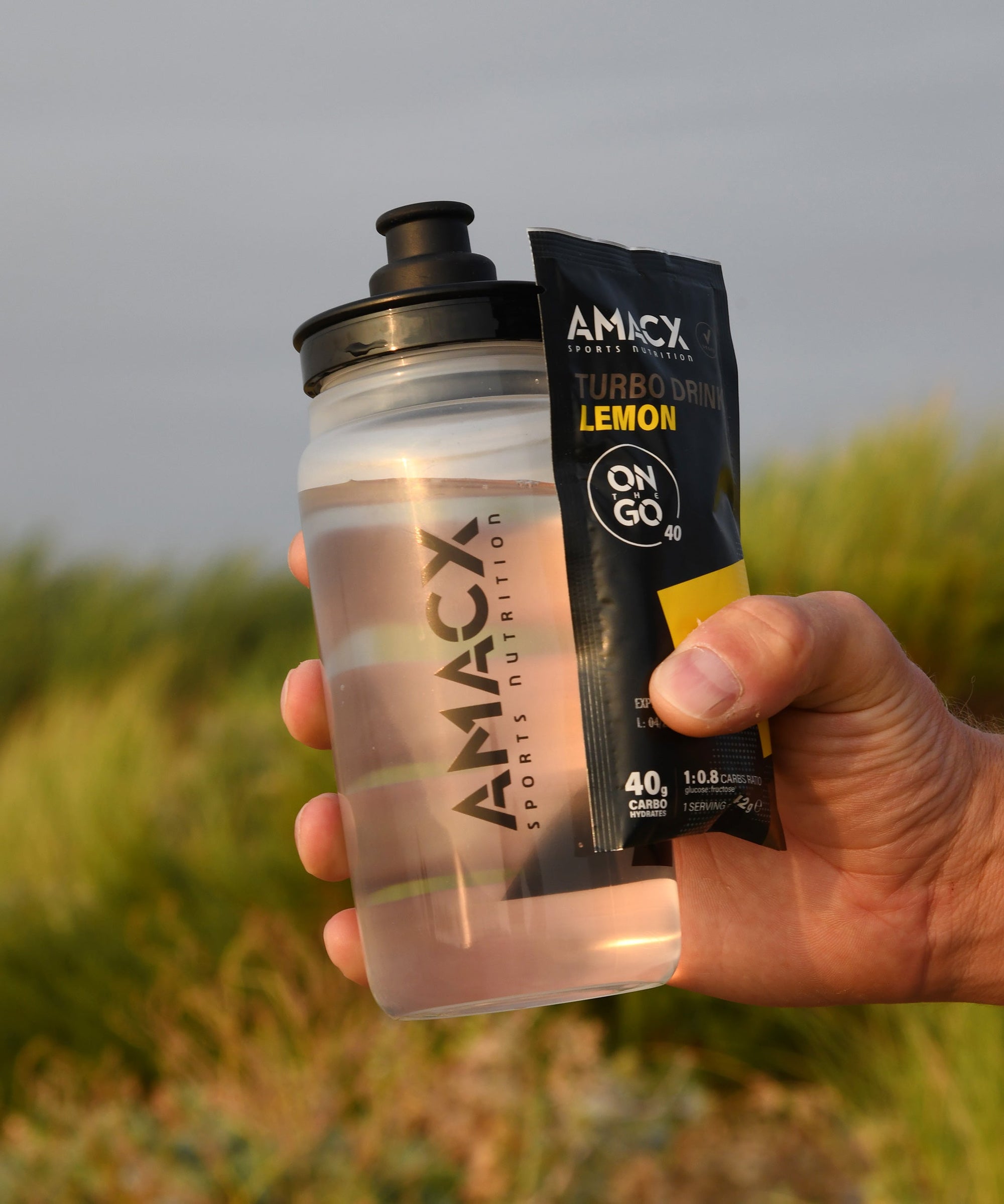 Hand holding Amacx Turbo Drink On The Go Lemon packet and clear sports bottle outdoors with blurred greenery background