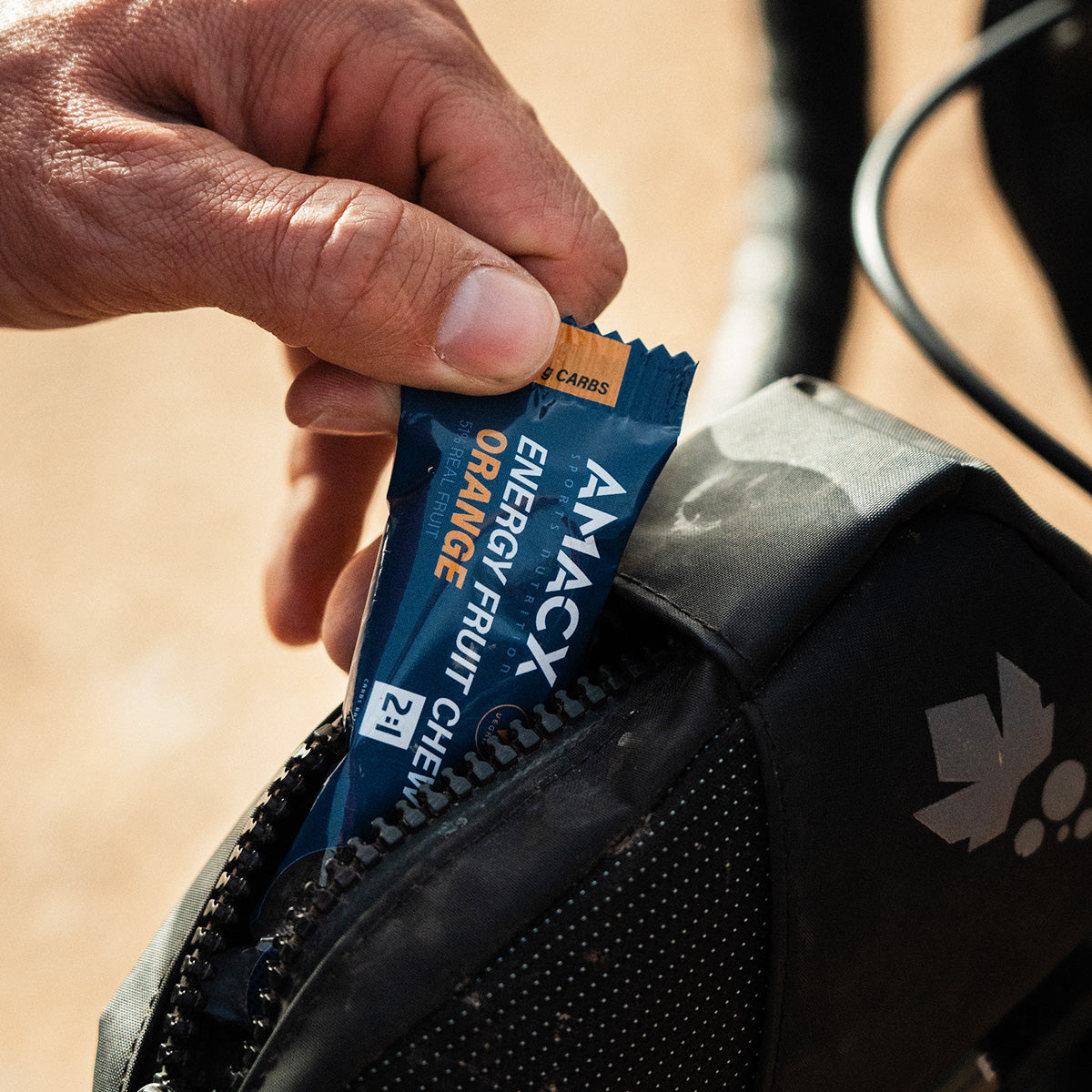 Hand placing Energy Fruit Chew Orange | 12 pack into black zippered bike bag in outdoor setting gallery position 4