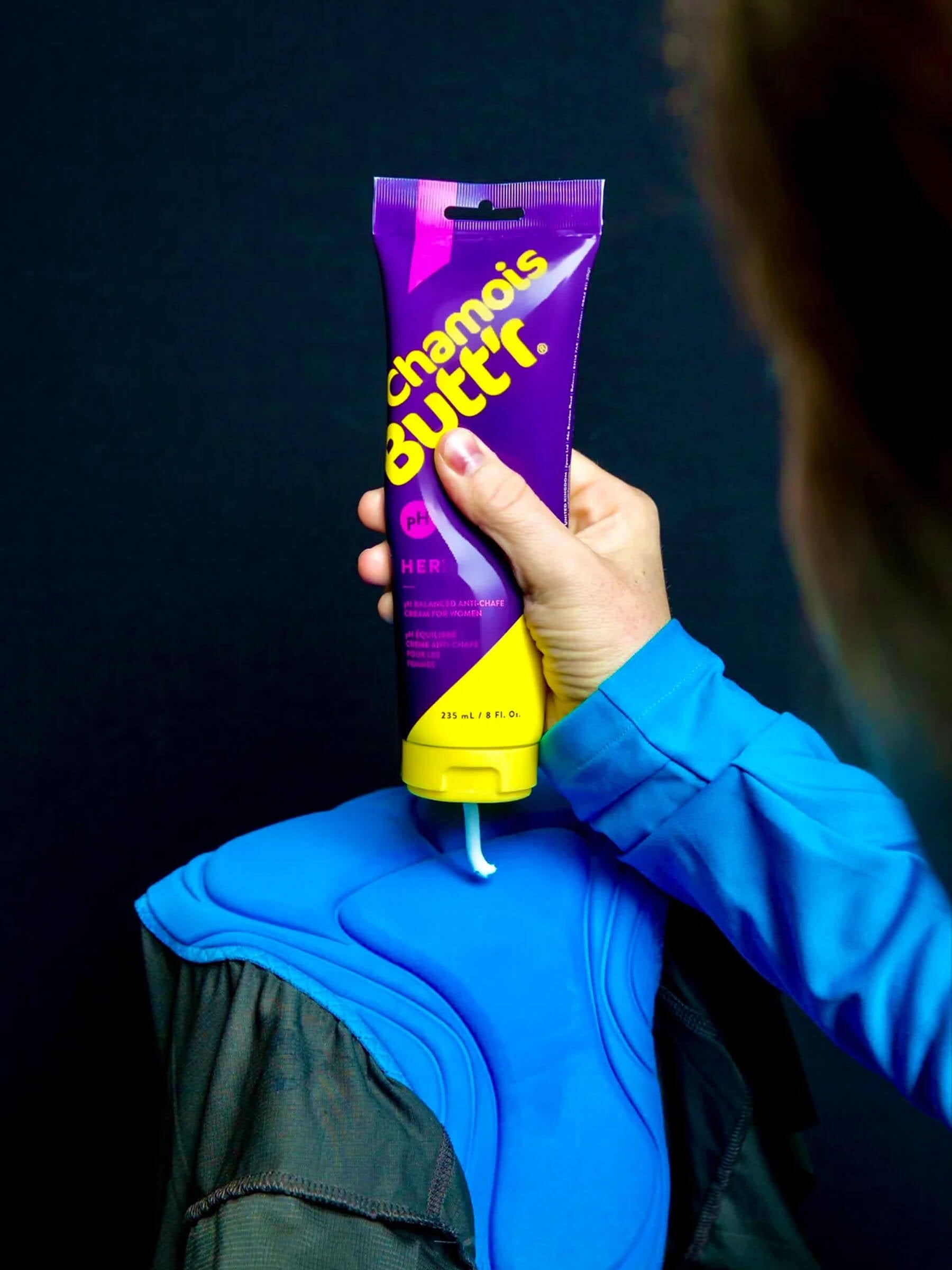 Person holding tube of Chamois Butt'r Chamois Cream - Women's dispensing cream onto blue cycling shorts padding