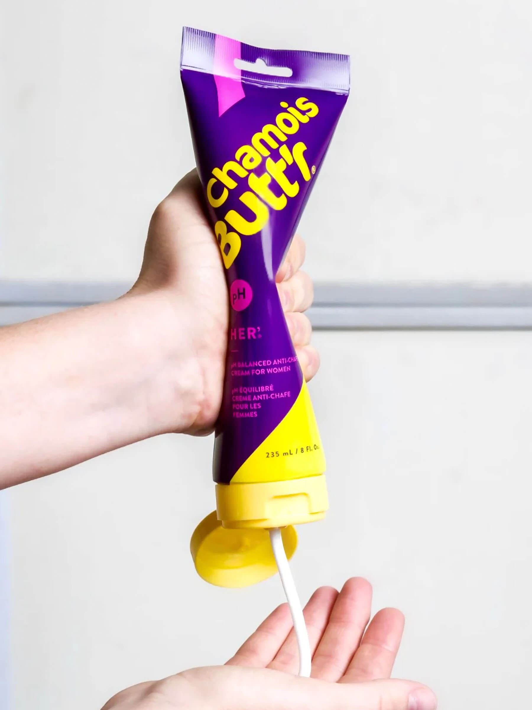 Hand holding and dispensing Chamois Butt'r Chamois Cream - Women's from purple and yellow tube against light background