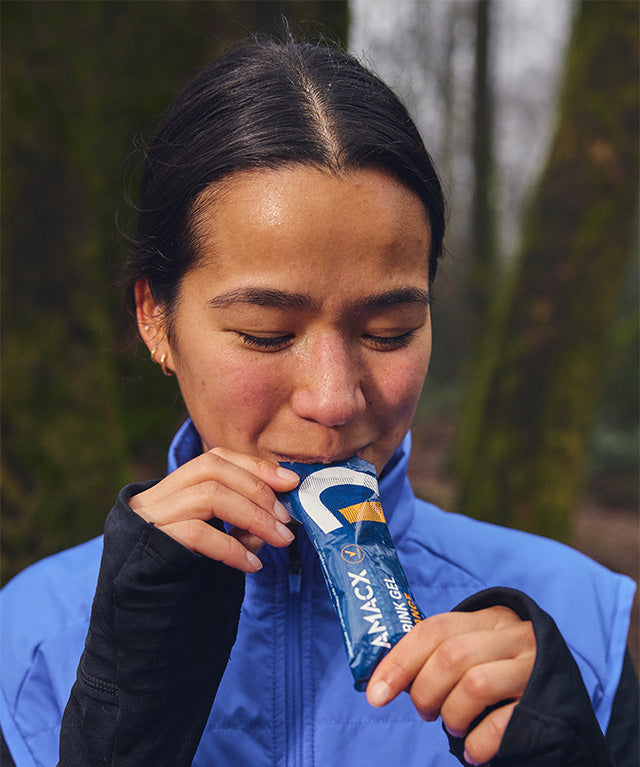 Woman in blue jacket outdoors consuming Drink Gel Orange | 12-pack from a blue sachet in portrait gallery position 4
