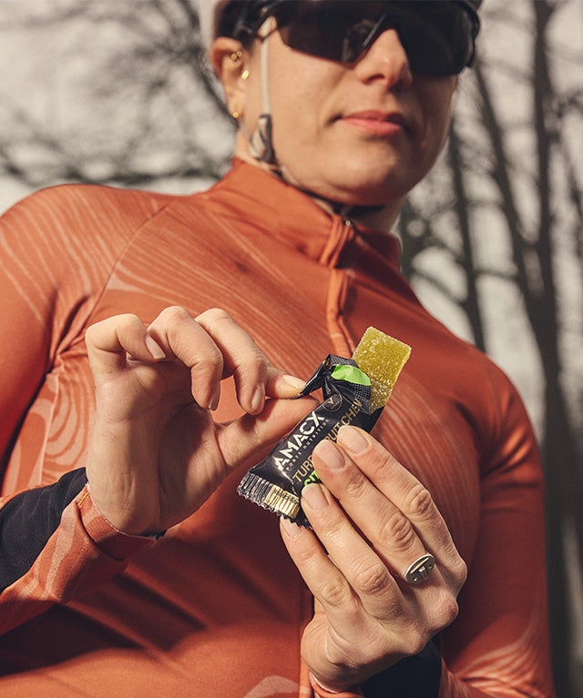 Person in outdoor gear holding and unwrapping Amacx Turbo Fruit Chew Citrus candy against a blurred natural background