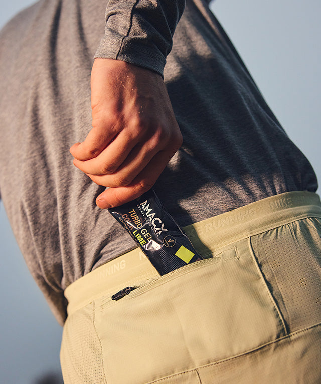 Person placing an Amacx Turbo Gel Cola Lime energy gel into the back pocket of light-colored athletic shorts.