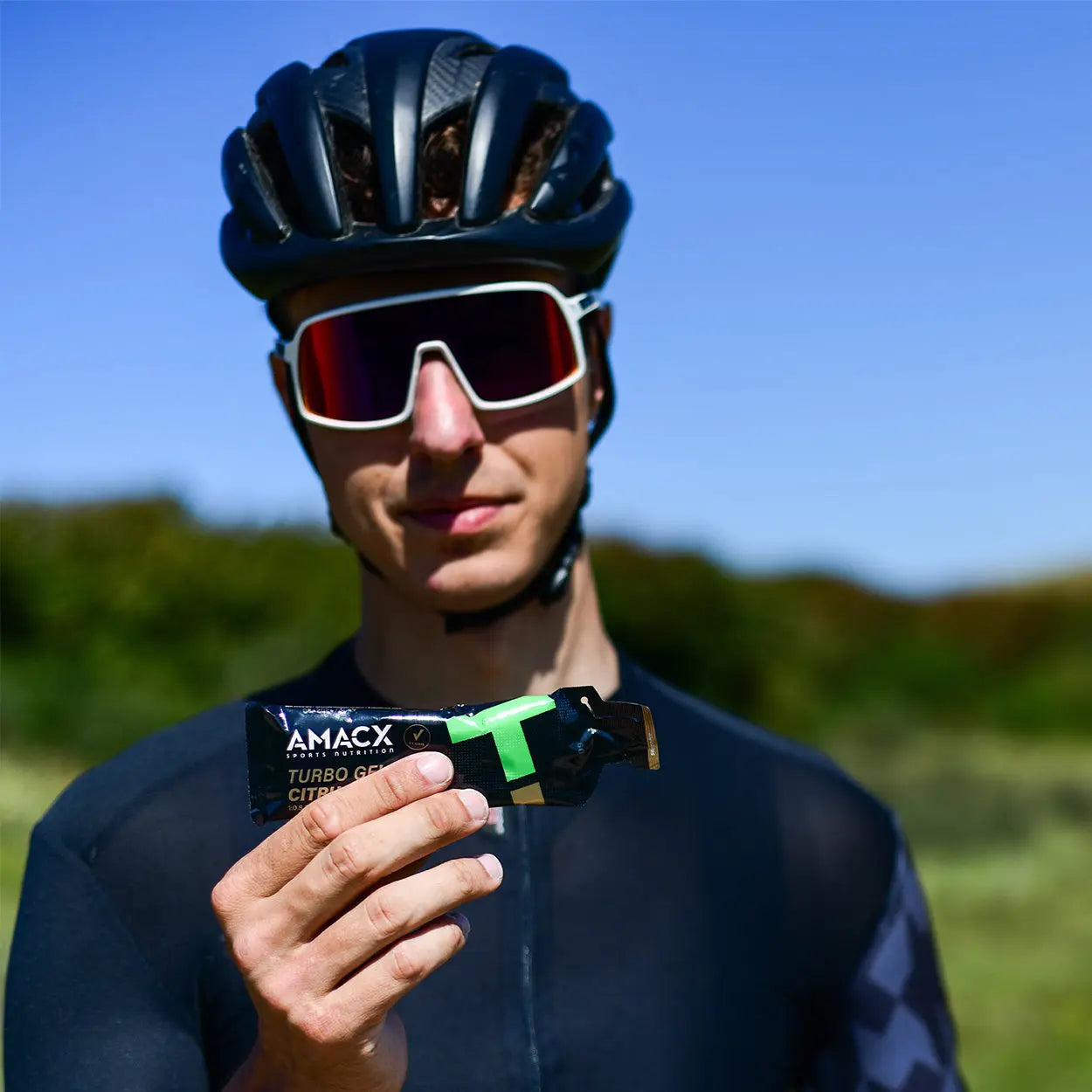 Athlete in cycling gear holding Amacx Turbo Gel Citrus energy gel outdoors with clear blue sky background.
