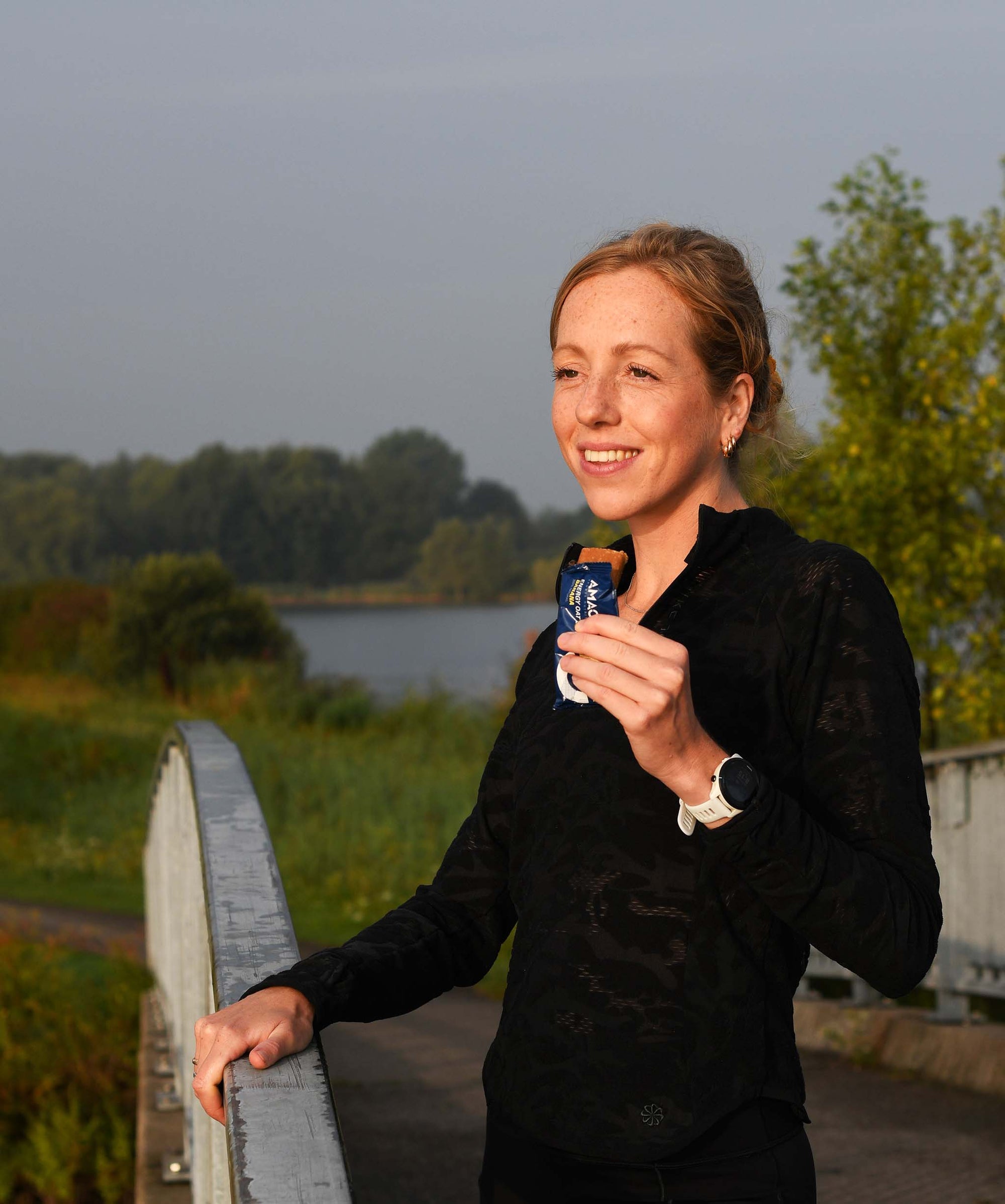 Woman enjoying an outdoor break holding an Amacx Energy Oat Bar Banana while standing on a bridge at sunrise
