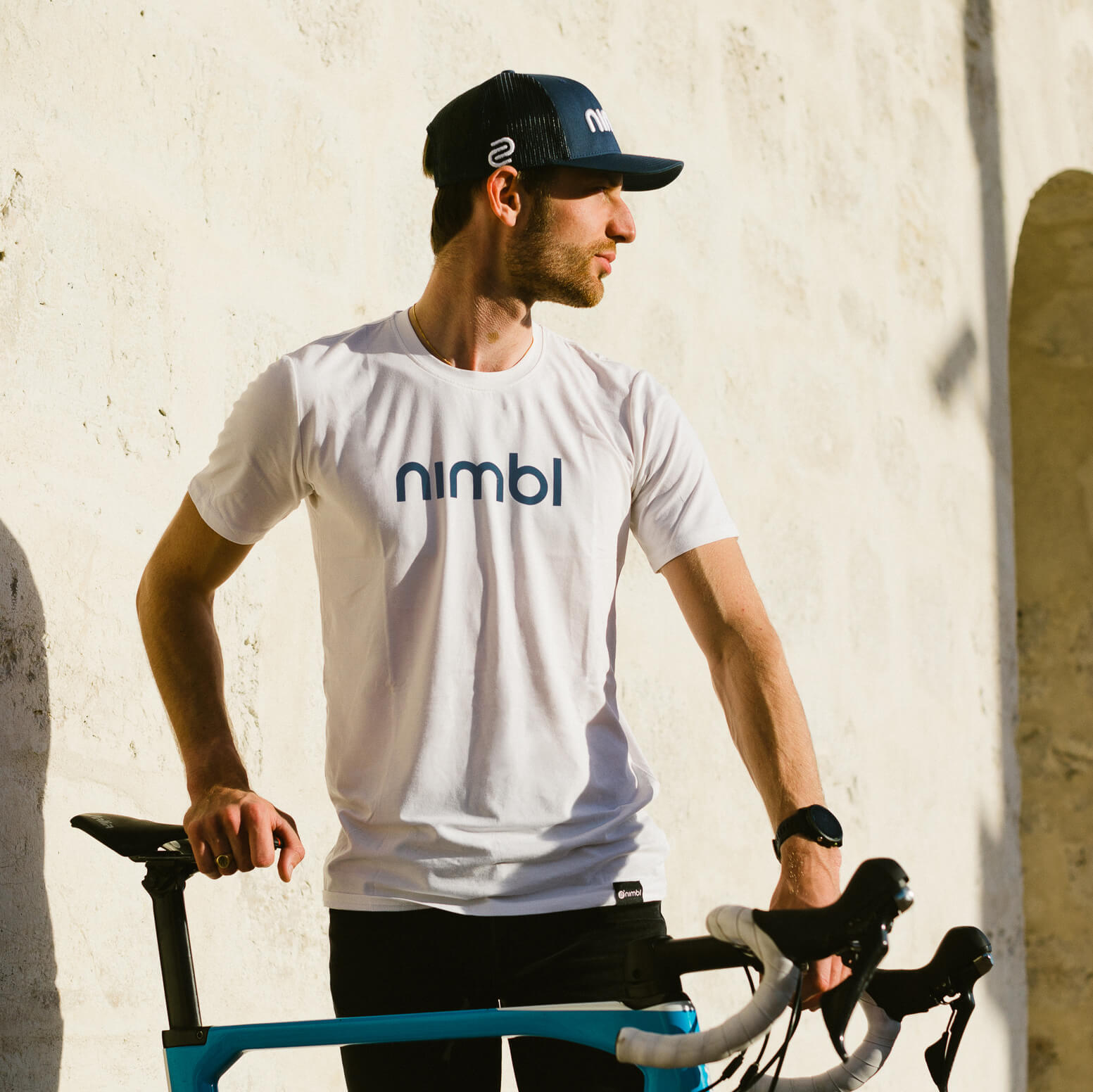 Man wearing Nimbl Cotton T-shirt - Men's standing next to blue bicycle in front of sunlit textured wall