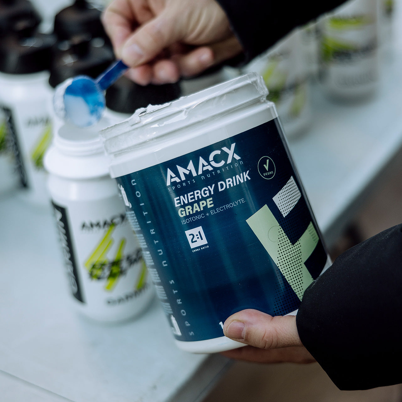 Hand holding a tub of Amacx Energy Drink | 1.0 kg with a blue scoop of powder near a shaker bottle