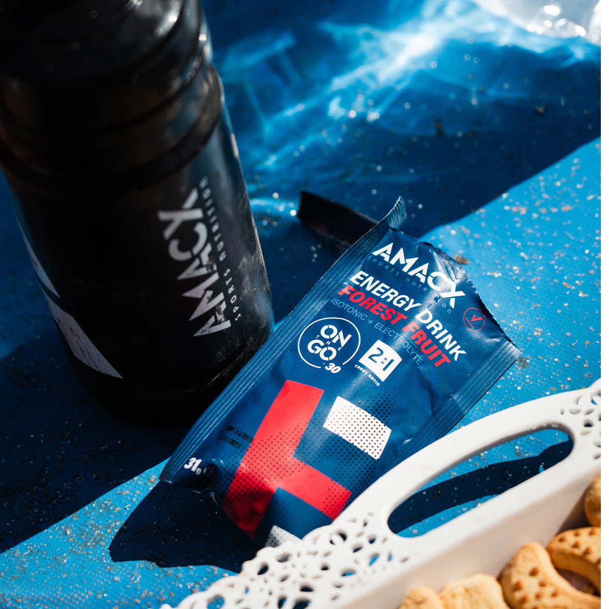 Amacx Energy Drink On The Go packet with black bottle and snacks on a blue surface in natural light