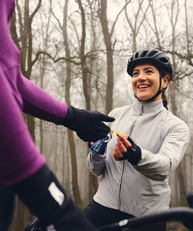 Woman in cycling gear smiling while sharing Amacx Energy Oat Bar Banana outdoors in forest setting